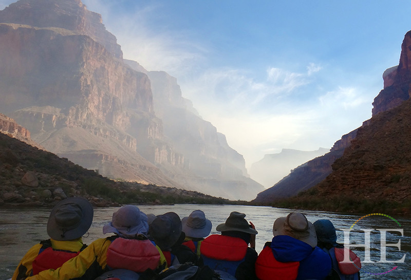 A group of travelers is on a raft on the Colorado River with red rocks in the distance on HE Travel's Splash Grand Canyon Rafting Tour.
