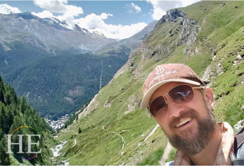 huge vistas of alpine mountains of switzerland, zach moses is in the foreground wearing hat, sunglasses, and beard