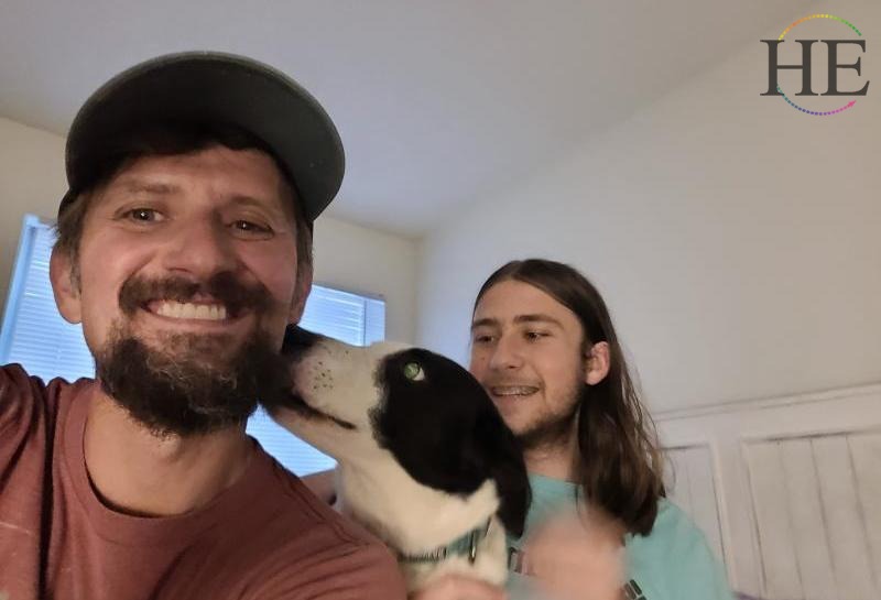 Zach Moses in a baseball hat with border collie Otzyl likcing zachs face and son Archimedes with long hair